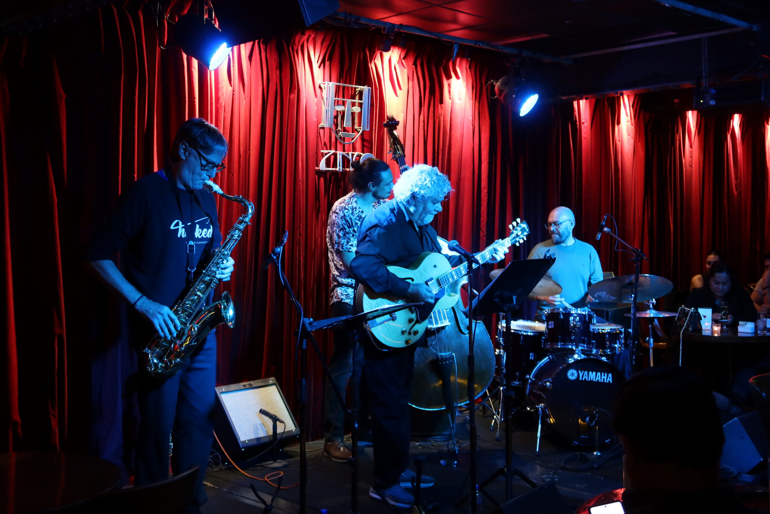 4 integrantes de un grupo de jazz tocando el saxofon, guitarra, chelo y bateria en el club de música jazz "Cinco"